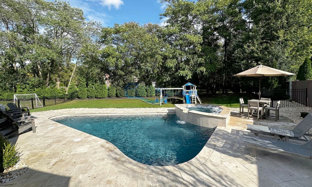 Backyard swimming pool with a slide, surrounded by lush greenery and a sunny sky, featuring a stone deck and lounge chairs for relaxation.